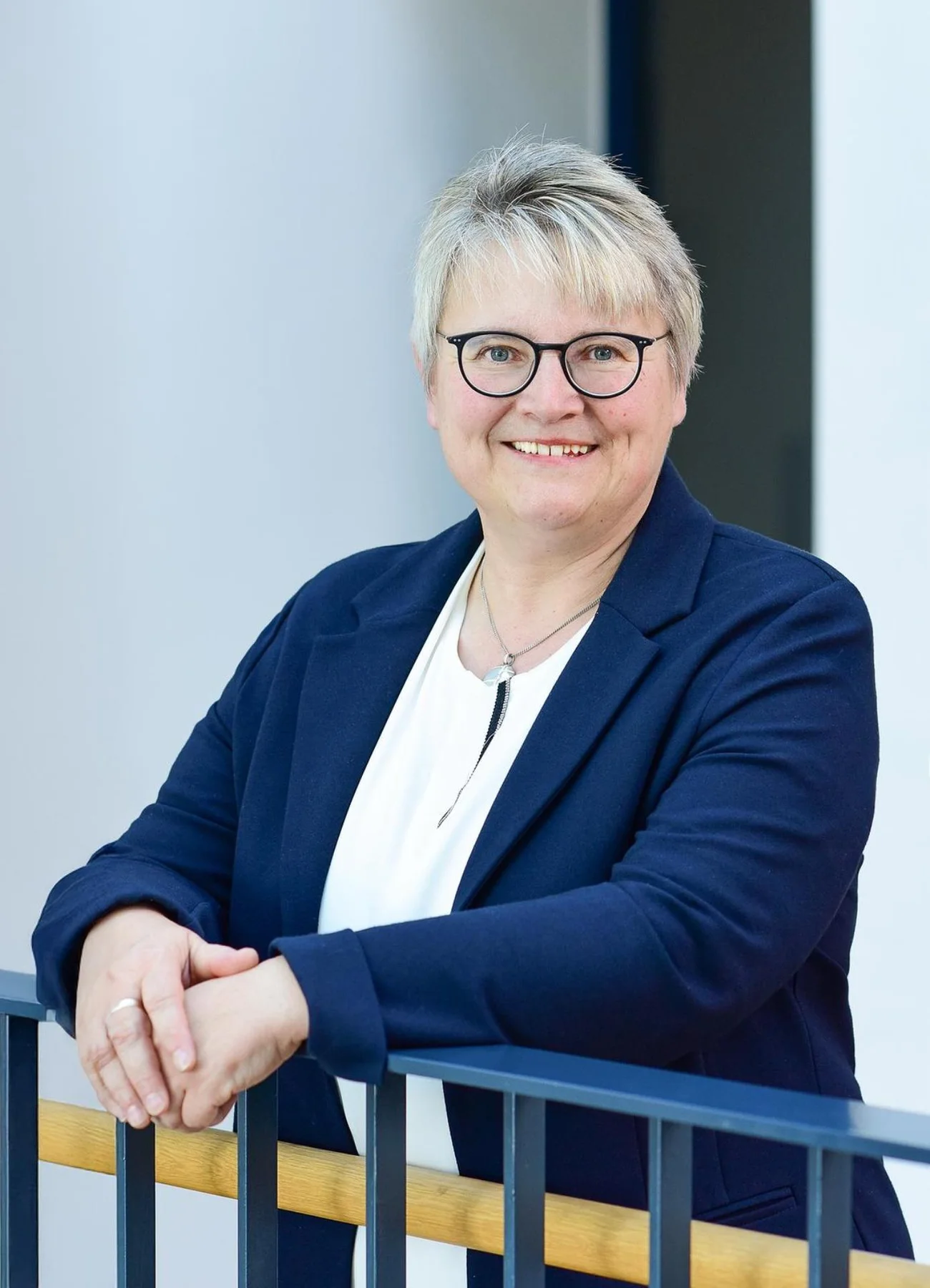 1. Bürgermeisterin Anita Ferber Stadt Monheim Porträt einer lächelnden Frau mit kurzen grauen Haaren, Brille, weißem Hemd und blauem Blazer, die an einem Geländer lehnt.
1. Bürgermeisterin der Stadt Monheim