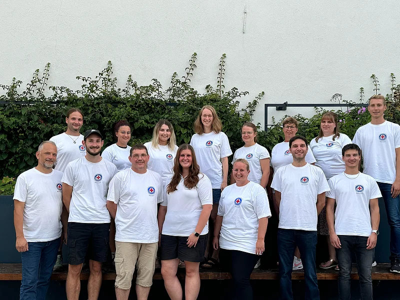 Gruppenfoto von 15 Personen in weißen T-Shirts mit dem Logo der Wasserwacht drauf, stehen blickend in die Kamera vor einer weißen Wand mit Pflanzen im Hintergrund.