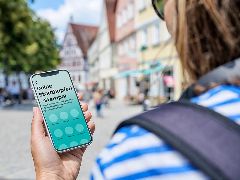 Mit der neuen Stadthupferl-App Städte erkunden und Stempel sammeln Person hält Smartphone mit App „Deine Stadthupferl-Stempel“ vor unscharfem Stadtplatz mit Fachwerkhäusern.