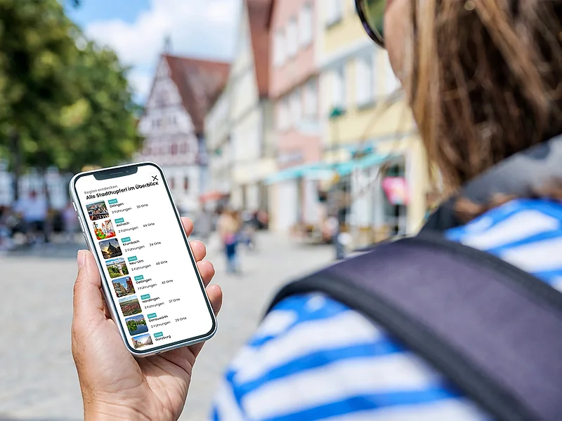 Neue Stadthupferl-App von Bayerisch Schwaben Person hält Smartphone mit App zur Stadttourenübersicht vor unscharfem Stadtbild mit Fachwerkhäusern.