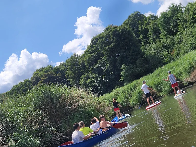 Sechs Personen paddeln in einem Kanu und drei Personen stehen auf Stand-Up-Paddle-Boards auf einem Fluss bei bewölktem Himmel.