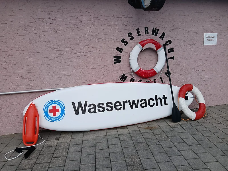 Rettungsbrett mit Aufschrift „Wasserwacht“ und Rettungsringen vor rosa Wand mit Uhr und Schild „Einspringen verboten“