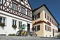 Tourist-Information der Stadt Monheim/Monheimer Alb: ein Fachwerkhaus mit drei Stockwerken, weißen Wänden und braunen Balken. Rechts davon ein gelbes zweistöckiges Gebäude mit roten Fensterläden. Zwei Personen mit Fahrrädern stehen davor und schauen eine Broschüre an.