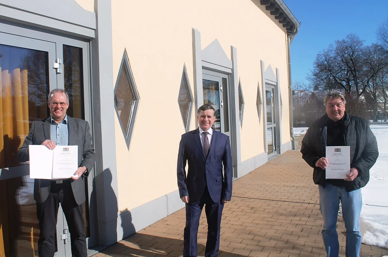 Dankurkunde für die beiden langjährigen, verdienten Stadträte Alfons Meier und Josef Steinhart Drei Männer stehen vor einem hellen Gebäude auf einem gepflastertem Platz, zwei halten Urkunden in den Händen, Schnee rechts im Bild liegt auf dem Boden.