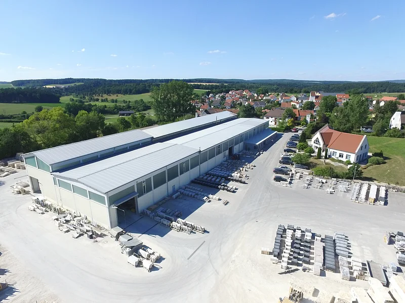 Natursteinwerk Böswald GmbH Luftaufnahme eines Industriegebäudes von einem Natursteinwerk mit asphaltierten Lagerflächen und Fahrzeugen darauf. Ein Dorf, grüne Landschaft und blauer Himmel im Hintergrund.