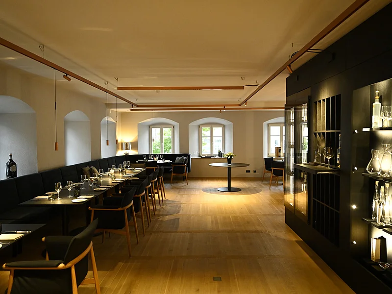 Innenansicht des Restaurants im Hotel by Ferber, in Monheim. Auf der rechten Seite ein großes schwarzes Regal mit verschiedenen Glasbehältern, Weingläsern und Flaschen. Auf der linken Seite schwarze Holztische mit schwarzen Sitzbänken und schwarzen Stühlen zu sehen.