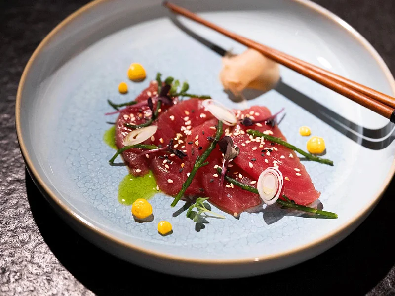 Dünne Scheiben rohen Fischs mit grünen Kräutern, Sesam, gelbem Mais und roten Zwiebeln auf einem schwarzen Tisch im blauen Teller mit Essstäbchen serviert.