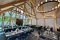 Großer Veranstaltungsraum einer Eventlocation mit offenem Dachstuhl und sichtbaren hellen Holzbalken oben an der Decke. In Reihen aufgestellte schwarze Tische mit denkelblauen Stühlen. Tische sind gedeckt mit Gläsern, Servietten, Besteck und Blumen in Vasen. Links im Bild große Glasfront ebenfalls mit hellen Balken. An der Decke große, runde, moderne Leuchten angebracht.