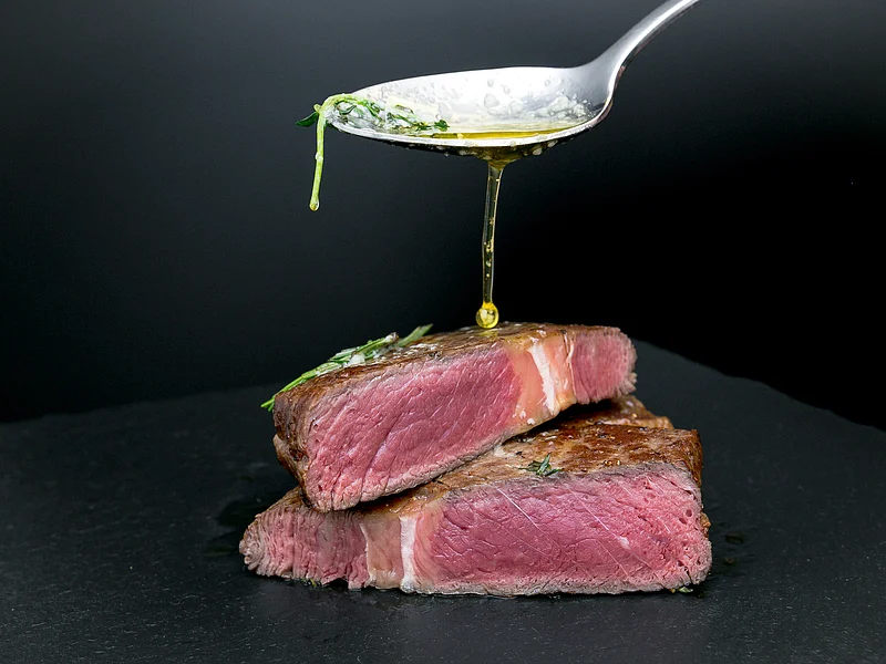 Steaks für Genießer im Hotel by Ferber - Hotel, Steakhouse, Bar in der historischen Altstadt von Monheim Ein Löffel gießt Öl über zwei aufeinander gestapelte, halbierte Rindersteaks auf einem schwarzen Hintergrund.