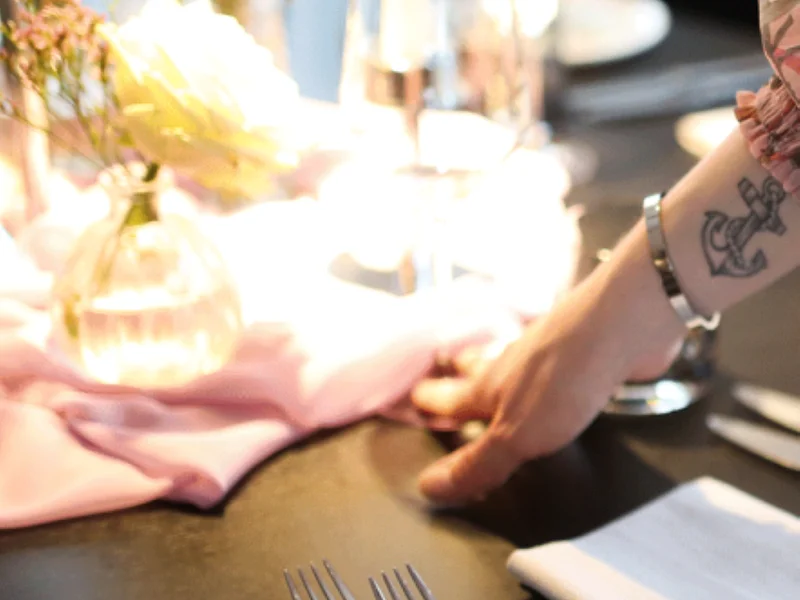 Hand einer Bedienungskraft an einem dekorierten Tisch mit zwei Gabeln und einem bunt bemaltem Teller mit Blumenmuster. Rechts davon eine weiße Serviette und unscharf erkennbar ein Weinglas mit einer weißen Kerze daneben.