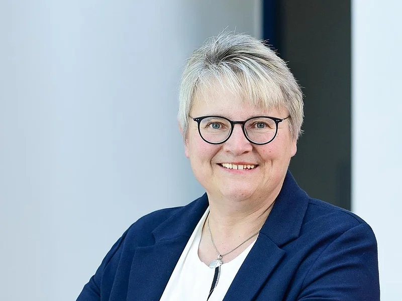 Porträt einer lächelnden Frau mit kurzen grauen Haaren, Brille, weißem Hemd und blauem Blazer, die an einem Geländer lehnt.
1. Bürgermeisterin der Stadt Monheim
