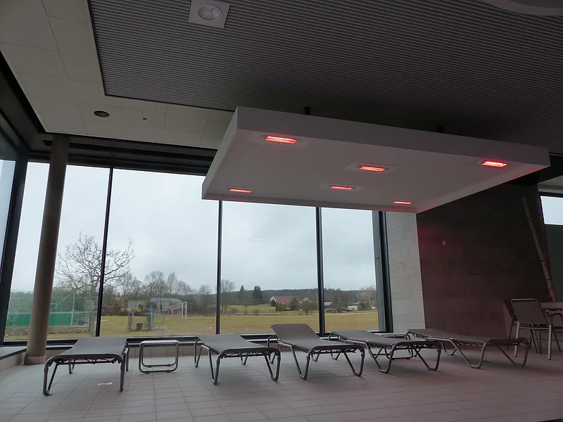 Innenraum eines Hallenbades mit Liegestühlen vor großen Fenstern und einer Deckenleuchte mit roten Lichtern.