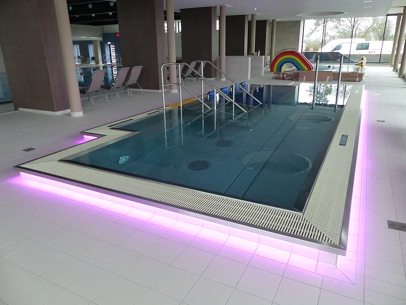 Innenansicht eines Schwimmbades, dem Monheimer Jurabad: ein mit Wasser befülltes Schwimmbecken mit Stufen und Handläufen, umgeben von Fliesenboden. Der Rand des Beckens ist außenrum rosa/lila beleuchtet. Im Hintergrund stehen drei Liegestühle und eine Regenbogenrutsche, sowie ein weiteres Becken.