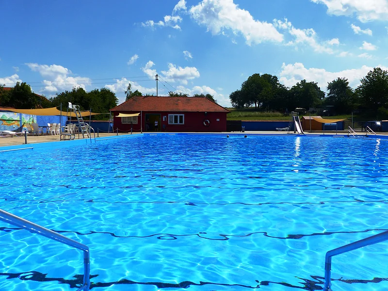 Das Freibad in Monheim: Schwimmbecken mit klarem Wasser, umgeben von Liegestühlen, einem roten Gebäude und Umkleidekabinen im Hintergrund.