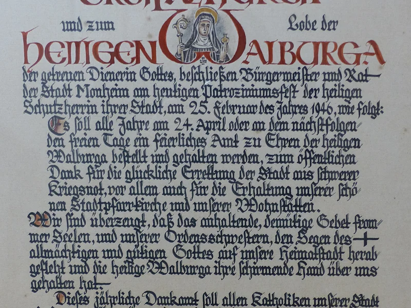 Dankamt-Urkunde von 1946 von Bürgermeister und Rat der Stadt Monheim Ein handgeschriebenes Dokument mit dekorativen roten und schwarzen Buchstaben, das eine religiöse Widmung enthält. Hierin gelobt der Bürgermeister sowie der Rat der Stadt Monheim, ein alljährliches Dankamt zu Ehren der Heiligen Walburga abhalten zu lassen, für die Verschonung der Stadt im Zweiten Weltkrieg.
