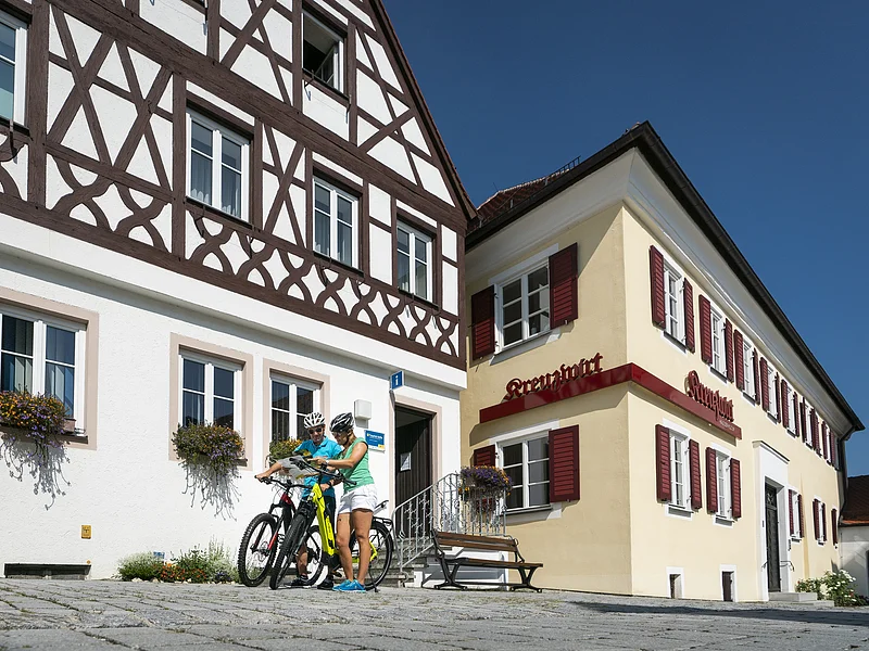 Tourist-Information der Stadt Monheim/Monheimer Alb - im Schindlerhaus Tourist-Information der Stadt Monheim/Monheimer Alb: ein Fachwerkhaus mit drei Stockwerken, weißen Wänden und braunen Balken. Rechts davon ein gelbes zweistöckiges Gebäude mit roten Fensterläden. Zwei Personen mit Fahrrädern stehen davor und schauen eine Broschüre an.