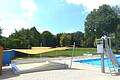 Außenbereich eines Freibads mit Planschbecken für Kinder, einem Schwimmerbecken, Rutsche aus Metall und gespannten Sonnensegeln bei Sonnenschein. Im Hintergrund grüne Wiese und Bäume.