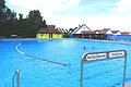 Ein Freibad mit einem großen Schwimmbecken, unterteilt in Nichtschwimmer- und Schwimmerbereich. Im Hintergrund sind Gebäude und Bäume zu sehen.