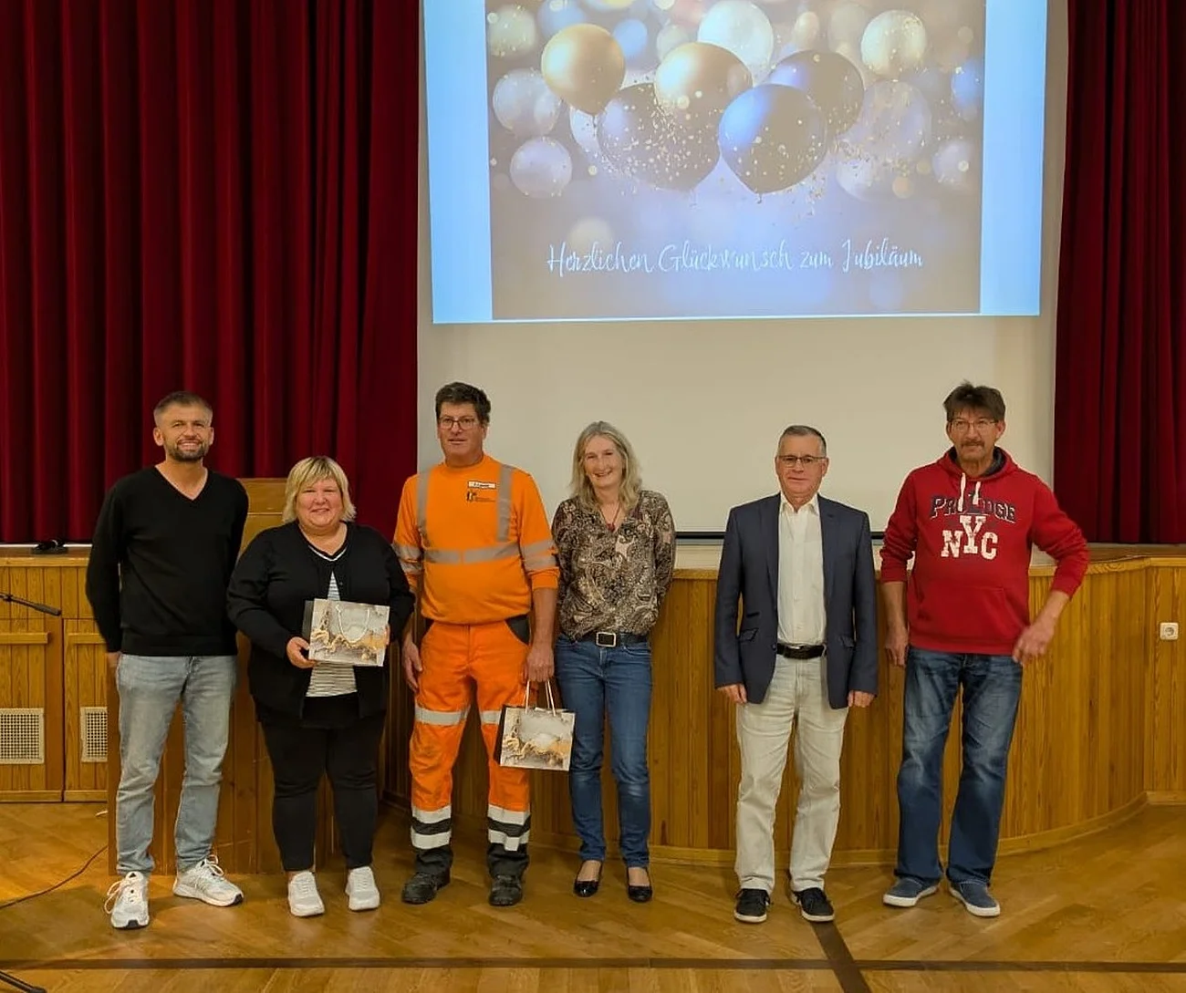Sechs Personen stehen nebeneinander auf einer Bühne vor rotem Vorhang und Leinwand mit Jubelbild und Text. Es handelt sich um den Personalrat der Stadt Monheim und geehrte Jubilare der Stadt Monheim.