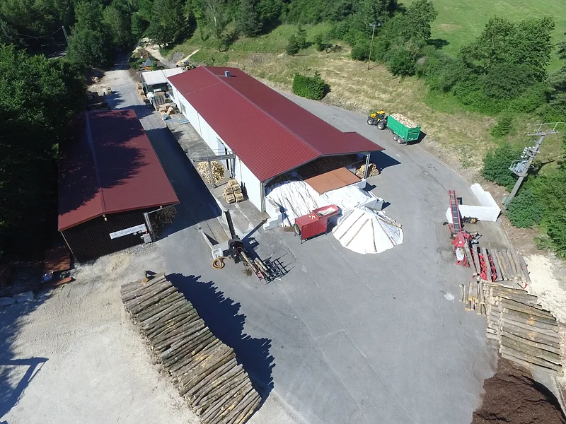 Forstunternehmen Steinle in Weilheim Luftaufnahme eines Holzlagers mit zwei großen Gebäuden, Holzstapeln und Maschinen auf Asphaltfläche, umgeben von Wiese und grünen Bäumen.