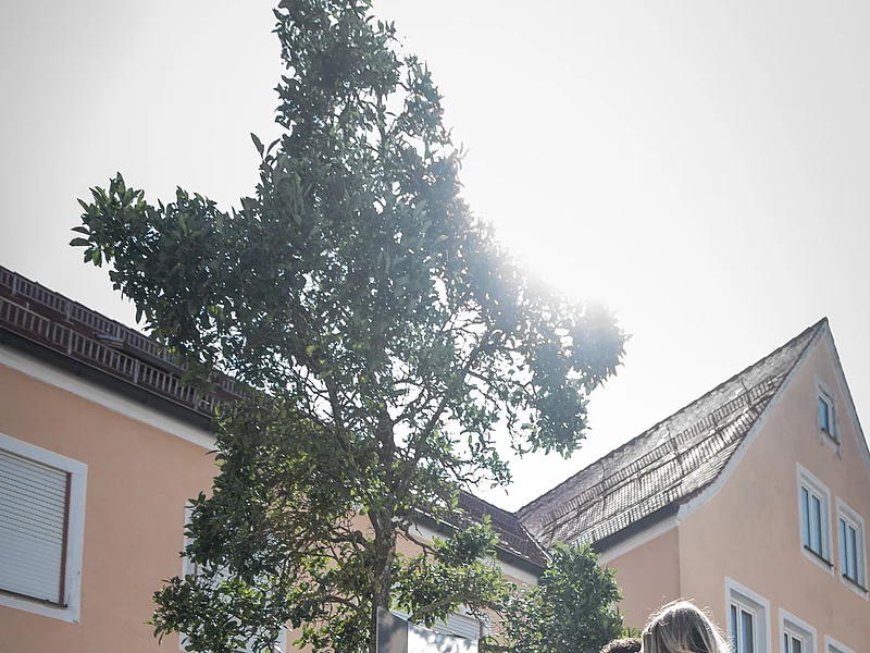 Zwei Personen stehen neben einer Steinskulptur des Buchstabens "N", daneben eine Informationstafel, beides steht auf einem Beet, mit einem Baum dahinter. Umgeben von einem gepflasterten Platz, Häusern und blauem Himmel im Hintergrund.