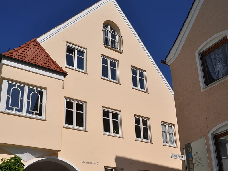 Buchstabenweg M-O-N-H-E-I-M Ein dreistöckiges Gebäude mit Giebel, einem Türbogen und mehreren Fenstern auf einem gepflastertem Platz. Vor dem Gebäude steht ein Informationsschild und ein großer grauer Buchstabe "M" aus Stein. Der Buchstabe ist ein Teil des Monheimer Buchstabenweges.