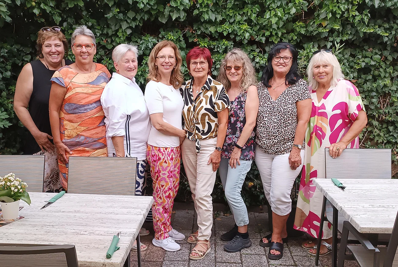 Neun Frauen stehen eng nebeneinander vor einer grünen Hecke, im Vordergrund Tische mit Stühlen und Besteck. Ehrenamtliches Helferteam Soziales Miteinander - 10 Jahre Fundgrube Monheim