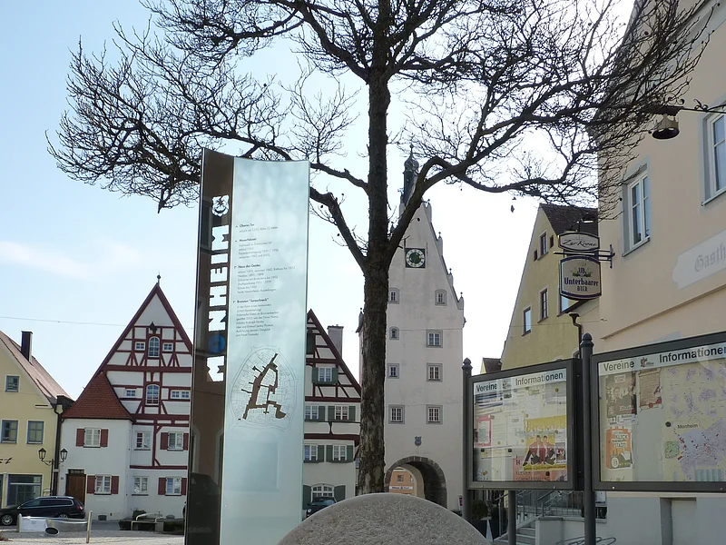 Im Vordergrund ein großer grauer Buchstabe "O" aus Stein mit einer Infotafel daneben auf einem gepflasterten Platz. Dahinter, rechts im Bild zwei weitere Informationsvitrinen der Stadt Monheim, umgeben von historischen Gebäuden im Hintergrund. Der Buchstabe ist ein Teil des Monheimer Buchstabenweges.