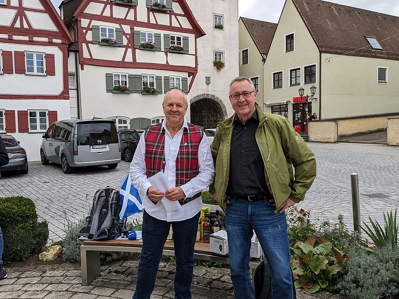 Zwei Männer stehen vor Fachwerkhäusern auf gepflastertem Platz und einer Bank mit Rucksack und Flaschen im Hintergrund. Die Bank ist umgeben von Pflanzen. Eine schottische Fahne - in weiß-blau - liegt auf der Sitzbank.