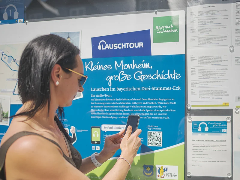 Frau mit Smartphone steht vor Infotafel zur Lauschtour in Monheim, Bayern, mit dem Titel "Kleines Monheim, große Geschichte" bei Sonnenschein. Frau hält ihr Smartphone an einen QR-Code.