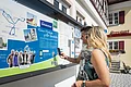 Startpunkt der Lauschtour "Kleines Monheim, große Geschichte"- an der Tourist-Information Monheim Zwei Personen vor einem weißen Gebäude, der Touristinformation Monheim betrachten eine Informationstafel mit einer Karte und Texten zur Lauschtour in Monheim und haben ein Smartphone in der Hand, mit dem sie einen Code abscannen.
