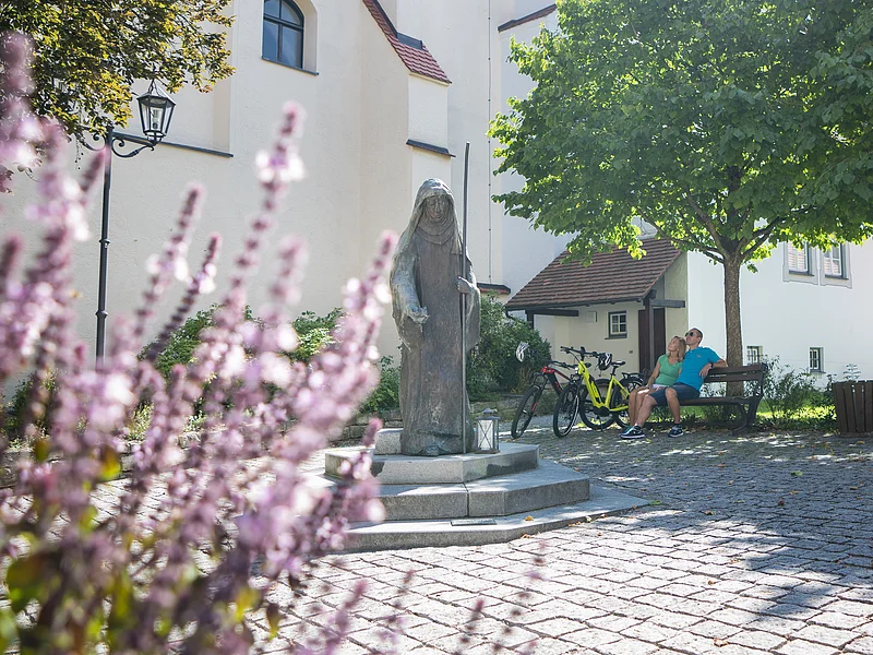 Steinfigur der heiligen Walburga auf einem gepflasterten Platz vor der Stadtpfarrkirche St. Walburga in Monheim, daneben zwei sitzende Personen auf einer Bank, dahinter ein Baum. Im Vordergrund unscharf Blumen.
