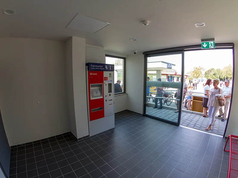 Wartehalle am Bahnhof Otting-Weilheim mit einem rot-grauem Fahrkartenautomat an der Wand, einer Glaseingangstür zum Außenbereich, einer roter Sitzbank rechts im Bild und gefliesten Boden. Vor der Tür im Außenbereich mehrere Personen bei Sonnenschein.