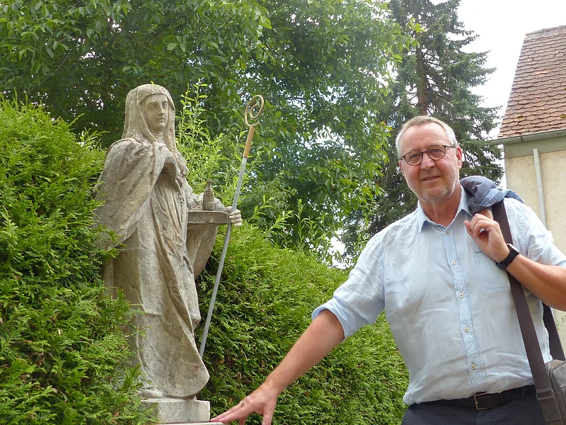 Eine Person mit Jacke über der Schulter steht neben einer Statue in einem Garten, umgeben von Büschen und einem Gebäude im Hintergrund.