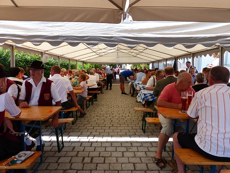 Dorferneuerung Flotzheim Mittagstisch Menschen sitzen unter einem großen weißen Zelt auf Bierbänken an Tischen bei einer Veranstaltung im Freien.