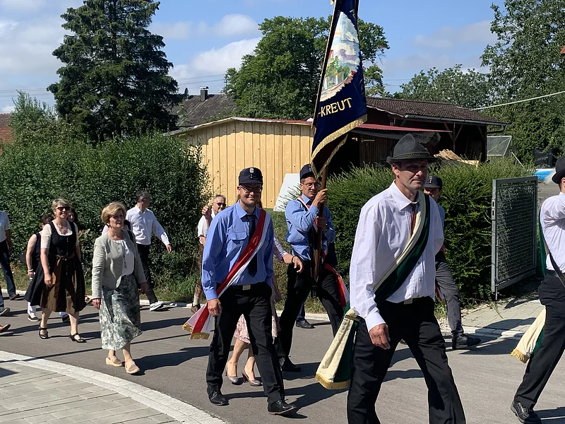 Dorferneuerung Flotzheim Festzug Gruppe von Menschen in traditioneller und formeller Kleidung bei einem Umzug im Freien an einem sonnigen Tag. Eine Person trägt eine große blaue Fahne mit dem Bild eines Ortes und mit dem Text "Kreut".