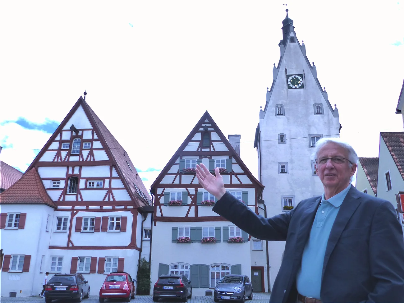 Der Monheimer Gästeführer Hanns Wenninger Eine Person mit Brille, im Sakko und blauem Hemd steht vor Fachwerkhäusern und einem Turm mit Uhr und zeigt darauf. Vor den Häusern stehen auf einer gepflasterten Fläche vier Autos geparkt.