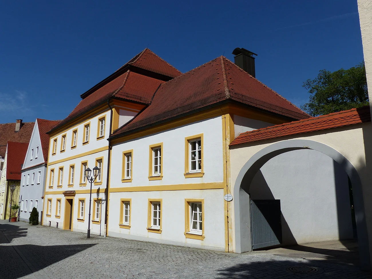 Pfarr- und Stadtbücherei Monheim Historisches weißes Gebäude, die Pfarr- und Stadtbücherei in Monheim, mit gelben Fensterrahmen und rotem Dach, daneben ein Torbogen auf gepflasterter Straße. Links vom Gebäude weitere Häuser in blau und grün.