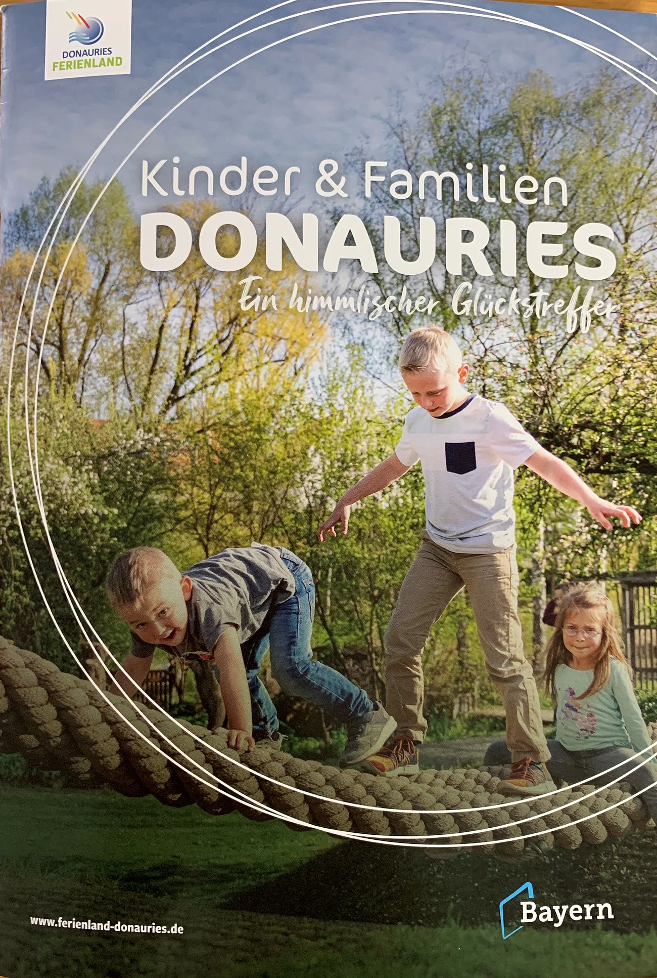 Broschüre Kinder & Familien DONAURIES Titelseite der Broschüre "Kinder & Familien Donauries" - Ein himmlischer Glückstreffer vom Ferienland Donau-Ries. Auf dem Titelbild drei Kinder spielend auf einem Kletterseil im Freien, umgeben von Bäumen und Wiese.