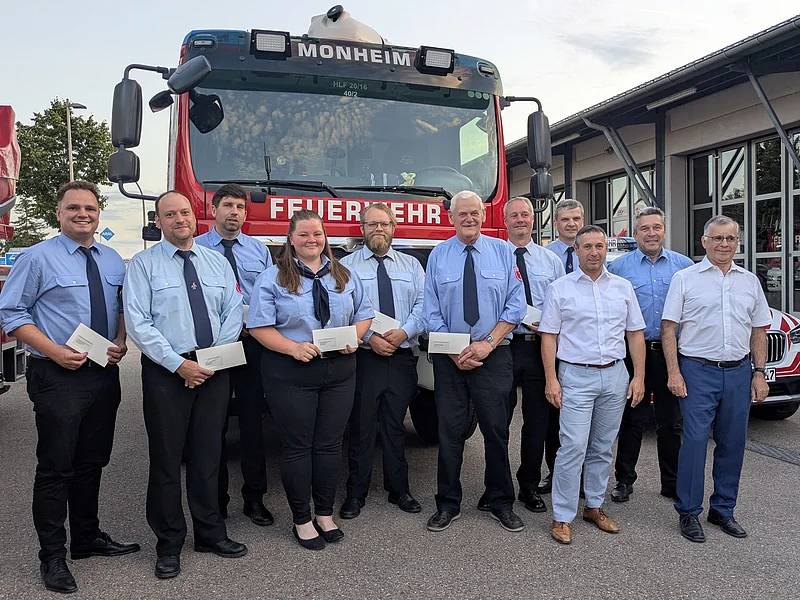 Abend der Feuerwehren - Spendenübergabe an die neun Feuerwehren Elf Personen in Feuerwehruniform und zwei Personen in Hemd stehen vor einem roten Feuerwehrfahrzeug mit der Aufschrift „Feuerwehr Monheim“ auf einem gepflastertem Platz.