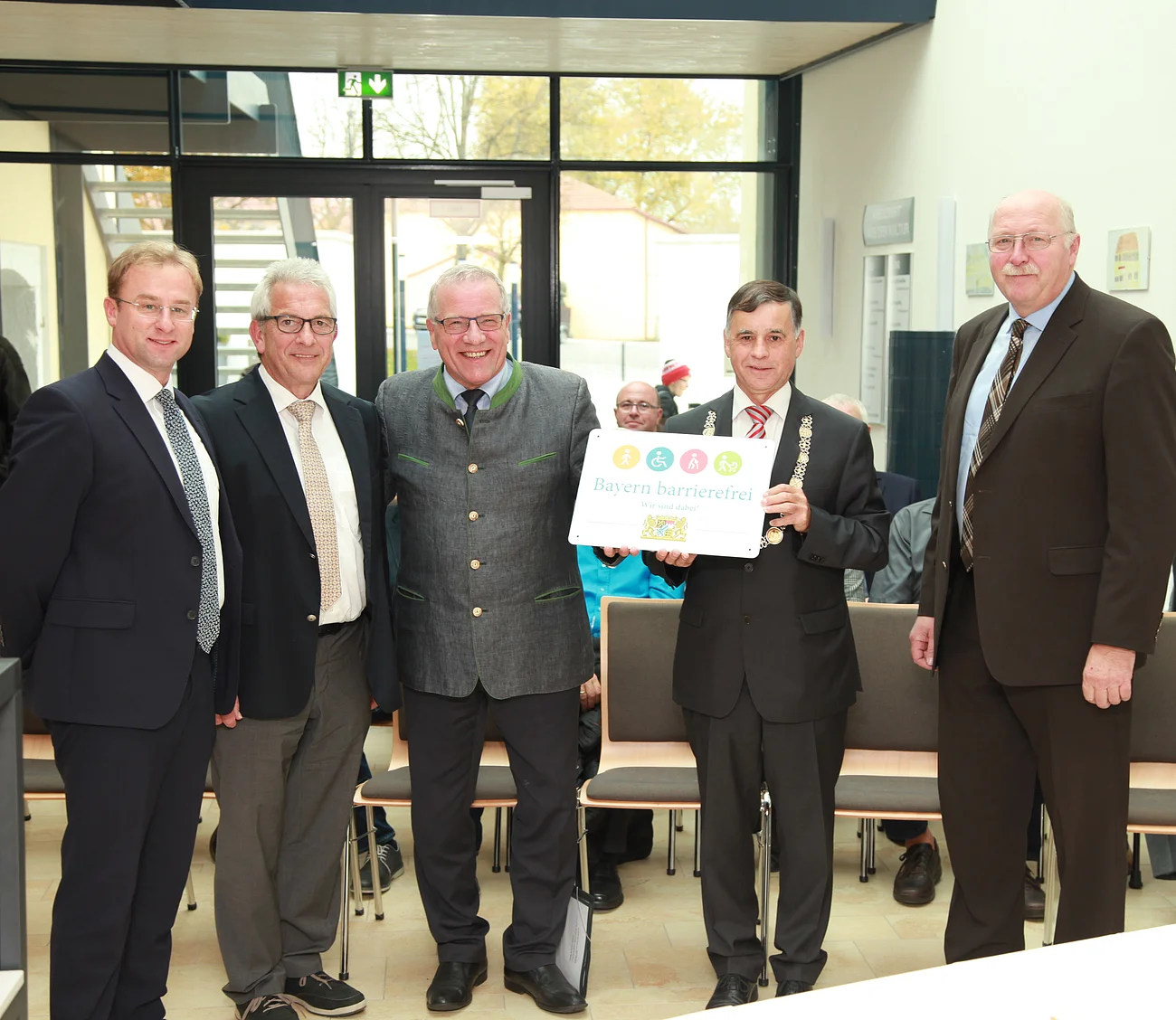 "Bayern barrierefrei" Fünf Männer in Anzügen stehen in einem Raum, einer hält ein Schild mit der Aufschrift "Bayern barrierefrei". Im Hintergrund große Eingangstür aus Glas.