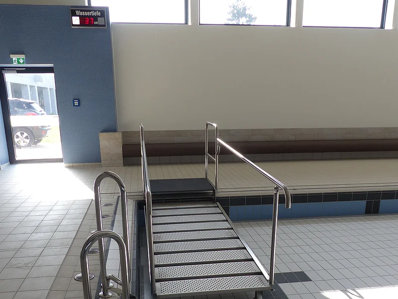 Barrierefreier Zugang im JURABAD Monheim Schwimmbeckenbereich in einem Hallenbad mit leerem Schwimmbecken mit Rampe aus Metall, die in das Becken führt und einer gefließten Treppe. Drei Fenster oben an der Wand und eine Ausgangstür links davon im Hintergrund.
