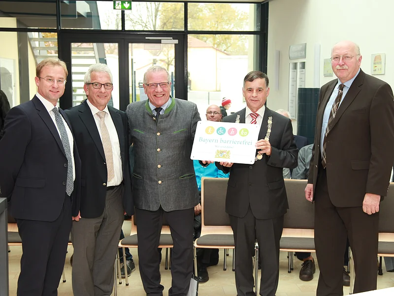 "Bayern barrierefrei" Fünf Männer in Anzügen stehen in einem Raum, einer hält ein Schild mit der Aufschrift "Bayern barrierefrei". Im Hintergrund große Eingangstür aus Glas.