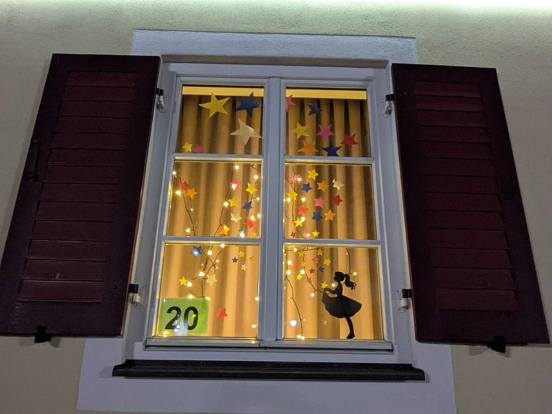 Adventskalenderfenster 21.12.2024 - städt. Kindergarten Adventskalenderfenster mit weißer Umrandung und geöffneten roten Fensterläden weihnachtlich dekoriert. Innen bunte Sterne umgeben von einer Lichterkette und unten rechts im Eck in schwarz die Silhouette eines Mädchens mit Kleid. Unten links im Eck die Zahl 20 auf Papier mit grünem Hintergrund.