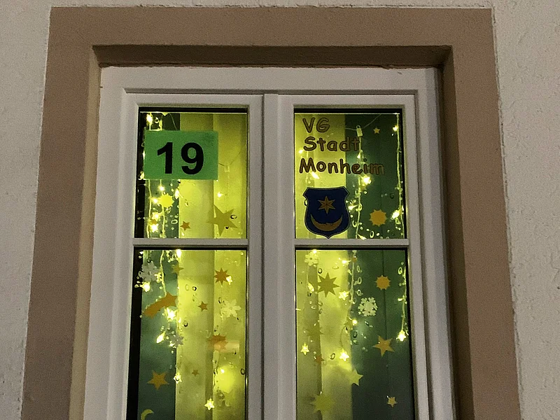 Adventskalenderfenster 19.12.2024 - VG & Stadt Monheim Adventskalenderfenster mit einer Umrandung in beige weihnachtlich dekoriert. Darin Beleuchtung mit Aufklebern von Sternen, Häusern und Bäumen. Oben rechts im Eck der Text „VG Stadt Monheim“ mit dem Stadtwappen der Stadt Monheim, sowie der Zahl 19 links davon auf Papier.
