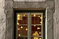 Adventskalenderfenster 14.12.2024 - Kath. Frauenbund Adventskalenderfenster mit grauem Rahmen außenrum weihnachtlich dekoriert. Darin Weihnachtsdeko wie Lichterketten, eine Weihnachtsmannfigur, weißer Tannenbaum mit Stern und von oben hängende Kugeln. Unten rechts im Eck die Zahl 14 auf grünem Hintergrund.