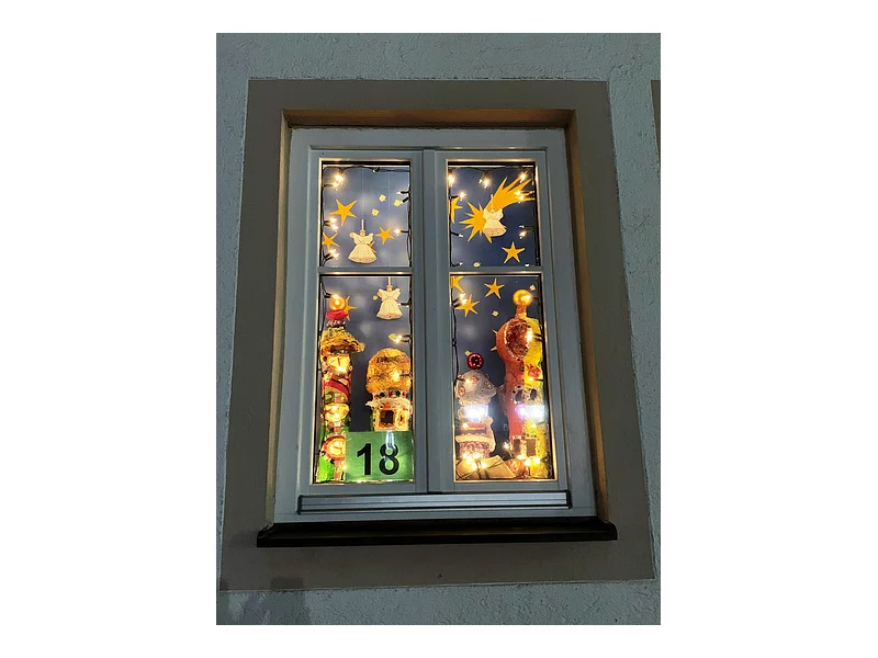 Adventskalenderfenster mit einer Umrandung in beige weihnachtlich dekoriert. Darin mit Weihnachtsbeleuchtung beleuchtete Engelsfiguren, eine Sternenschnuppe und Sternen. Unten links die Zahl 18 auf grünem Hintergrund.