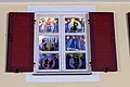 Adventskalenderfenster 10.12.2024 - Gailachia Adventskalenderfenster mit weißen Rahmen und roten Fensterläden weihnachtlich dekoriert. Bunte von einer Lichterkette beleuchtete Bilder mit blauem Vorhang dahinter, Christbaumkugeln und dekorativer Schnee hängen herunter. Links oben im Fenster das Logo des Faschingsvereins Gailachia Monheim.
