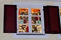 Adventskalenderfenster 01.12.2024 - Kolping Adventskalender mit weißem Rahmen und geöffneten roten Fensterläden weihnachtlich dekoriert. Innen ist Weihnachtsbeleuchtung und Dekoration wie rote Christbaumkugeln, Sterne oben und rote Geschenkschleifen. In der Mitte des Fensters rechts ist die Zahl 1 auf Papier angebracht. Darunter ein Portrait eines Mannes zu sehen.