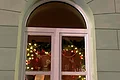 Adventskalenderfenster 03.12.2024 - Stadtkapelle Adventskalenderfenster mit Weihnachtsbeleuchtung, von oben hängenden roten Kugeln, Lebkuchenfiguren, Musikinstrumenten und einem Plüschelefanten mit einer weißen Mütze im Inneren.